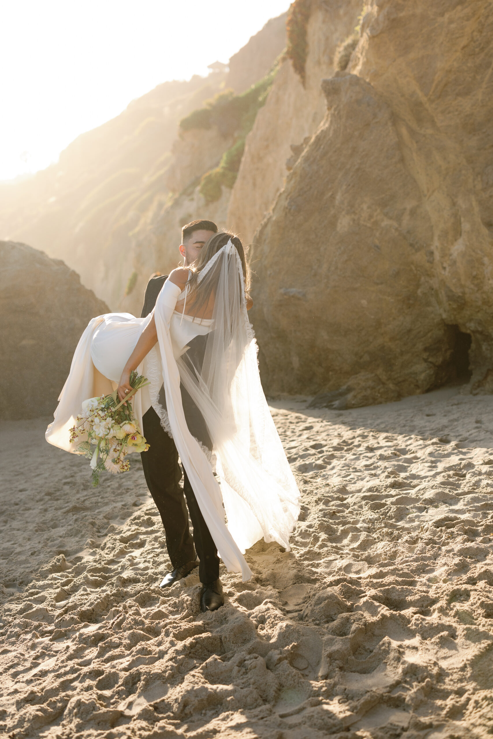 Bride and groom kissing on a sandy beach beneath coastal cliffs at sunset, captured for a luxury destination wedding venue feature
